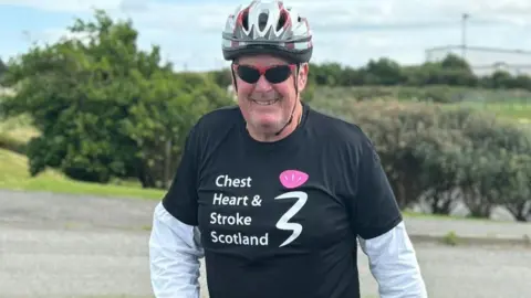 Scottish Rugby Ken MacAulay is wearing a black t-shirt  over a white cycling shirt. The shirt has the words "Chest, heart and stroke" printed on the chest. He is smiling and wearing a cycling helmet and sunglasses