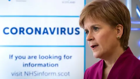 PA Media Nicola Sturgeon, with light brown hair and wearing a purple top, stands to the right of shot in front of a coronavirus sign 