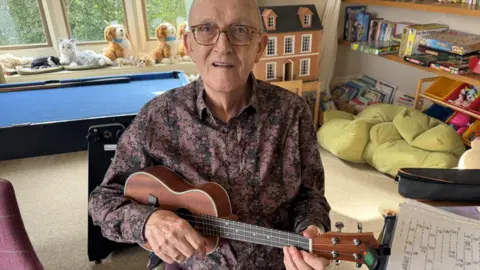 An elderly man sat holding a ukelele wearing a purple patterned shirt and wearing glasses. Behid him a doll's house and books and a bean bag