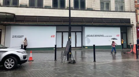 Pete Cooper/BBC Outside view of the H&M from across the road. The building is boarded up with white signs that have H&M in red written on them.