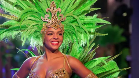 Thursford Christmas Spectacular A female dancer in a gold-coloured costume is wearing an intricate, golden tiara with large green feathers flowing from it.