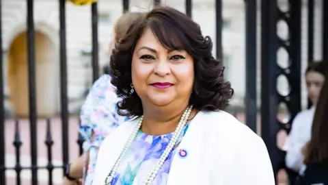 A head-and-shoulders photo of a woman with shoulder-lenght curly brown hair smiling in front of the black gates of Buckingham Palace. She's wearing a flowery dress, a white blazer, and a pearl necklace.