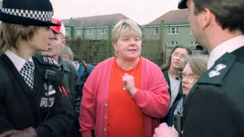 Margaret Haney photographed in 1997 outside a house on Stirling's infamous Raploch estate, with two police officers to either side during a 'paedophile-hunter' raid