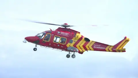 A Cornwall Air Ambulance helicopter flying in the sky. It is red and yellow and has the black and white Cornish flag on it.