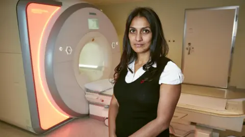 British Heart Foundation A woman standing in front of an MRI scanner which has orange light around its outer frame. She is wearing a black top.