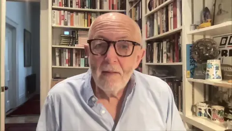 BBC International editor Jeremy Bowen wearing pale blue shirt and sitting in front of bookshelf
