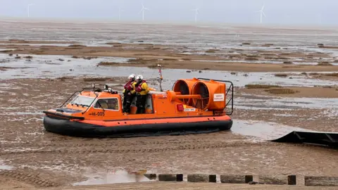 An orange-coloured hovercraft on Hoylake beach with wind turbines in the background. There are two men on the hovercraft.