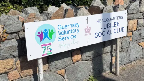 Granite wall with bushes in the background with a white sign displaying the Guernsey voluntary service logo.