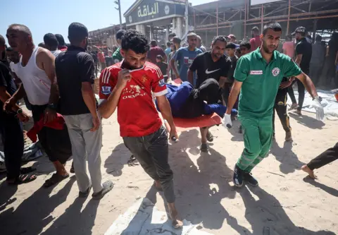 NurPhoto via Getty Images Several men carry an unidentified injured person on a stretcher outside the cafe with a crowd of people all around and blue sky in the background.