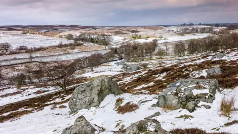 Danielrao/iStock/Getty Images Snow on rocks, trees and moorland in the North York Moors. A winding road can be seen in the background, and the sky is full of clouds.