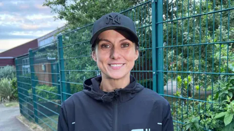 Kate Bradbrook/BBC Women in her thirties wearing a black baseball cap and black running jacket. She is standing in front of green fencing surrounding a school. She is smiling and looking directly at the camera. 