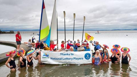 A group of swimmers in Clevedon marine lake wearing colourful umbrella hats. They are holding a banner that says 'thanks to National Lottery Players'
