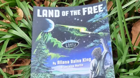 Lorraine Martin The book Land of the Free lies on grass. The cover shows a young man with long hair standing, looking up at the night sky with his right arm raised in the air. The moon can be seen in the top left corner, while a mysterious green light tracks across the sky.