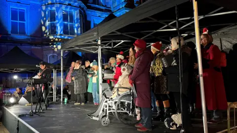 A group of people on a stage in front of an illuminated building. Some are wearing Christmas hats.