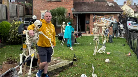 A man in a yellow hoodie and grey shorts standing in a garden with lots of skeletons and other Halloween decorations in it.