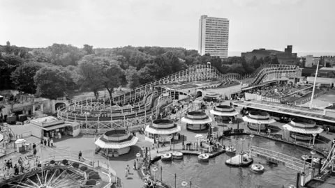 Mirrorpix via Getty Images A theme park as seen from the air, in black and white. Visible are a large rollercoaster and a pond with round boats on it. Trees and a large tower block can be seen behind the park.
