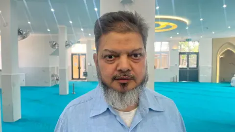 Moin Uddin, a man with a beard, wearing a blue shirt, stands alone in the same prayer room.