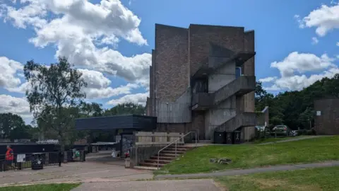 A brown-greyish building is surrounded by green trees and grass. The sky is clear and cloudy. Near the building, there is a small shop. 