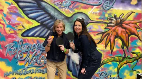 Former Traitors contestant Amanda Collier and radio presenter Allison Ferns pictured at the Brighton Table Tennis Club. They are stood in front of a canvas which says 'Solidarity - Refugees Welcome'. There is a hummingbird and flower painted onto the canvas. Amanda is wearing a shirt and cream trousers. Allison is wearing a black outfit. Both are holding table tennis bats.
