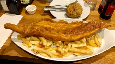 Stephen Peel A huge piece of battered fish sits on top of a plate of chips on a table. A pea fritter is on another plate above it.
