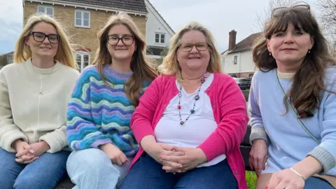 Jackie, wearing a pink cardigan and white top, sits next to her three daughters on a bench, with her house in the background 