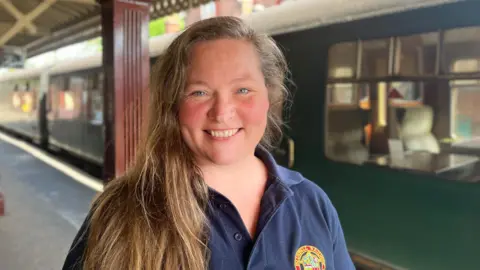 BBC/Josh McLaughlin A woman with long brown hair stood in front of a green train. 