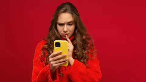 Getty Images A woman in a red jumper looks quizzical as she stares at her yellow mobile phone.