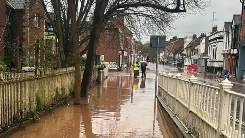 Flooding in Tenbury Wells in November, 2024. Brown floodwater on a pavement between two white fences. The street with shops in the background is also flooded. There is a small group of people in the distance. One is wearing a yellow high-visibility jacket and the other is holding an umbrella. 