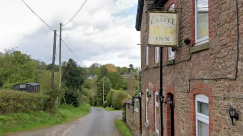 Google A narrow country road runs past a stone building with a hanging Castle Inn sign, surrounded by trees and hedges.