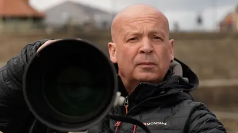 BBC Owen Humphreys is holding his camera and taking a photo. The angle is such that it is like we are looking down the lens. Humphreys is looking off into the distance, picking his shot. He is bald and has a black earring in one ear. He is wearing a black puffer jacket. The coast wall behind him is out of focus.