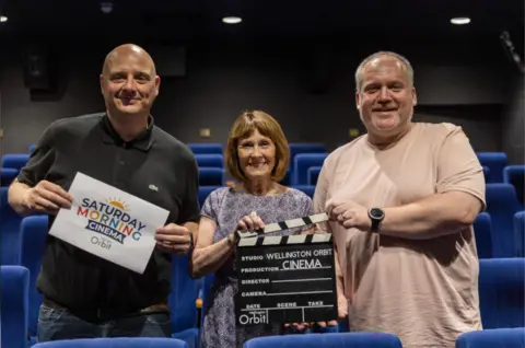 Telford and Wrekin Council A man in a black t-shirt holds a sign saying 'Saturday Morning Cinema'. A petite lady with brown hair holds a clapperboard saying 'Wellington Orbit cinema'. A man with a neutral colour t-shirt and a black watch hold the top of the clapperboard. They are all smiling at the camera. 
