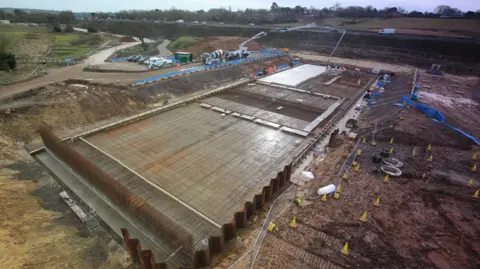 A construction site next to a motorway. There is a cement mixer, a crane, and construction workers, next to a huge metal centre point.