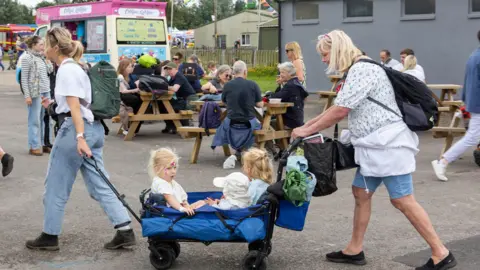 A Gorman Photography Two women, either side of a blue four-wheeled cart, pull the cart which contains three young children. A number of picnic benches with people sitting on them and an ice cream van appear in the background.