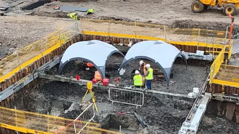 An archaeological dig has dug up a square area of earth a few metres below ground level with two canopies on the inside, a yellow barrier around its side and several workers in white helmets and yellow and orange high vis seen working on the site.