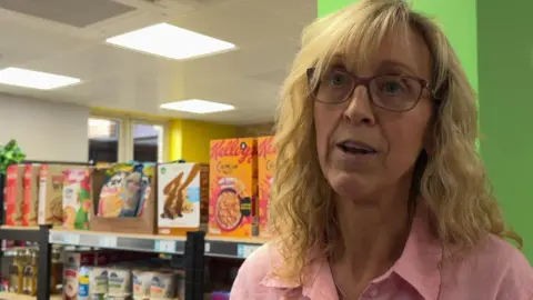 Claire Starr/BBC Kathryn Cliffe is standing in a community grocery with shelves of products behind her. She is wearing a pink shirt and has long blonde hair and glasses.