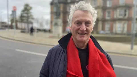 A man with grey hair, wearing a blue jacket and red scarf is stood outside a building. He is smiling at the camera