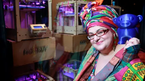 Camila Batmanghelidjh, the founder and chief executive of Kids Company, wearing colourful clothes and a colourful headdress. She is holding a 3D printed toy at the launch of Print Happiness, a pop-up store in London's Soho.