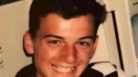 Ben West Sam West is wearing a polo shirt and has dark brown hair. He is sat in front of a white background 