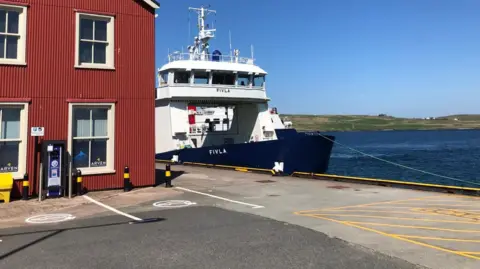 Part of the harbour area in Lerwick. A small ferry is stationary, next to a red building with cream windows. The sea and sky are blue and there's a green island in the distance. 
