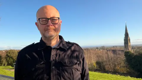 A bald man with a short grey beard is standing on a green hill. He is wearing a blue top with a dark jacket on top. Behind him is a number of trees and a church spire in the distance.