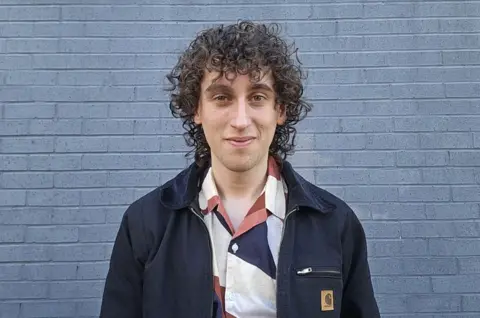 Khai Doak A man with curly brown hair in a red, white and navy top with a navy jacket stands in front of a grey brick walls and smiles at the camera