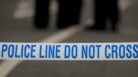 Blue and white tape carrying the instruction "police line do not cross" in capital letters. Out of focus in the distance are the legs of two or three people who are wearing dark trousers.