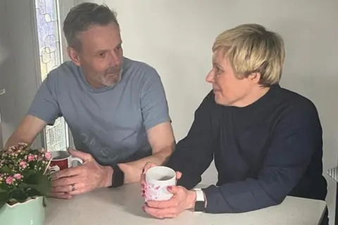 A woman on the right with short blonde hair next to a man with greying hair. They are both sat at a breakfast bar with cups of tea in their hands looking at each other.