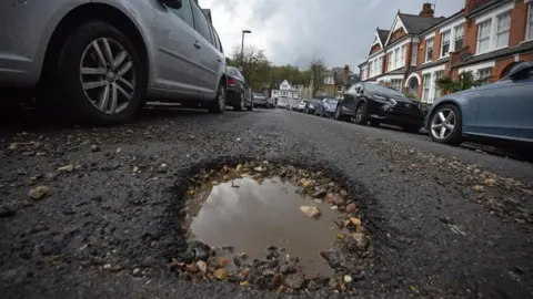 A pot hole filled with rain water and pebbles is in the foreground of a grey road on a suburban street with parked cars on either side of the road outside brick houses.