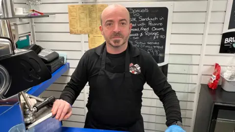 BBC/RICHARD EDWARDS Omar Musu - who runs Brushchetta cafe-bar in Filey - and is concerned the council tax premium could have "repercussions" for jobs - is standing behind the counter at his business. His right hand is on the coffee machine. There is a 'specials' menu board behind him. He is wearing a black long-sleeved T-shirt and a black apron.