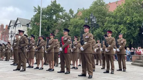 Soilders dressed in brown unifroms stand in formation, with crowds watching.