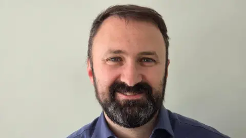 Ashley Comley A man with dark hair and a beard, wearing a blue shirt with an open collar. He smiles at the camera - it is a head and shoulders shot.