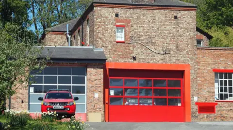 YNYCA A fire station with an engine parked behind a red garage door. A fire service car is parked in front.