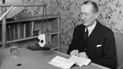 Black and white image of a man sitting at a desk with a folded piece of paper in front of him and a glass of water and a panel of knobs and an old looking microphone. In the background can be seen a book case of books and floral wall paper