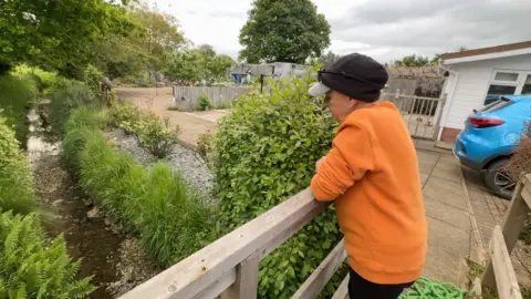 A lady stands on a bridge overlooking a stream that runs past her house. There is shrubbery on either side.